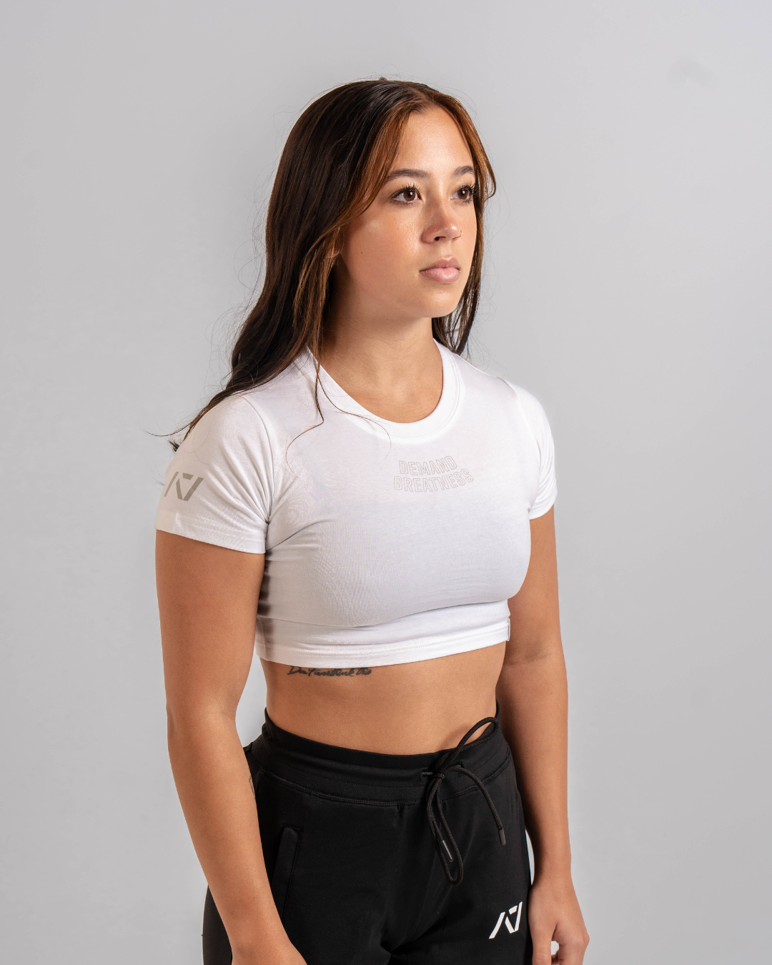 A woman wearing a white crop top with a Demand Greatness design on the upper chest and A7 logo on the upper back and sleeves.
