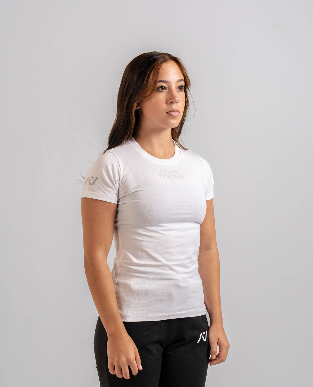 A woman wearing a white Demand Greatness True-Fit women's meet shirt with a logo on the upper chest and upper back, standing against a grey background.