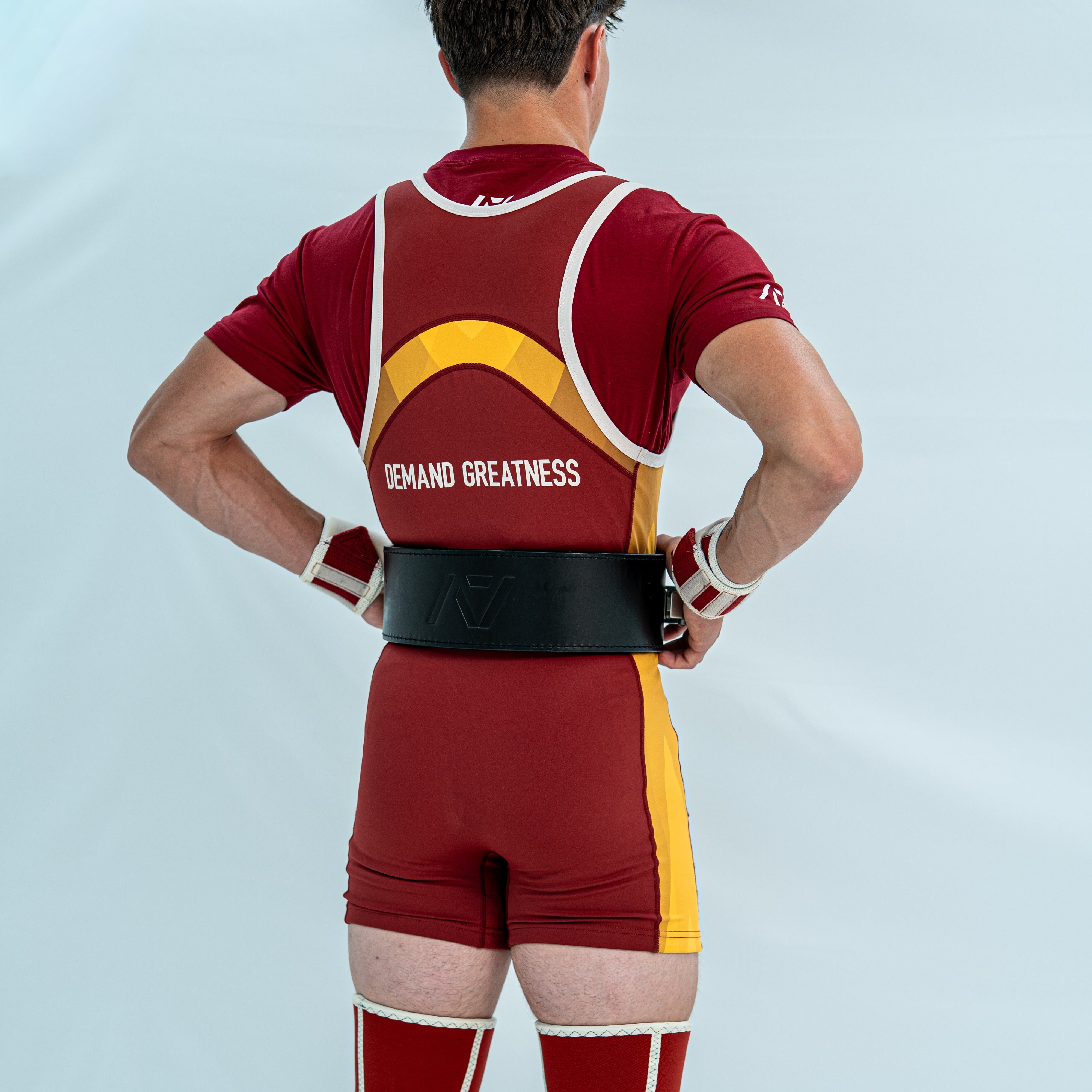Male model wearing a red and yellow powerlifting singlet with 'Demand Greatness' on the back of the singlet on a light grey background