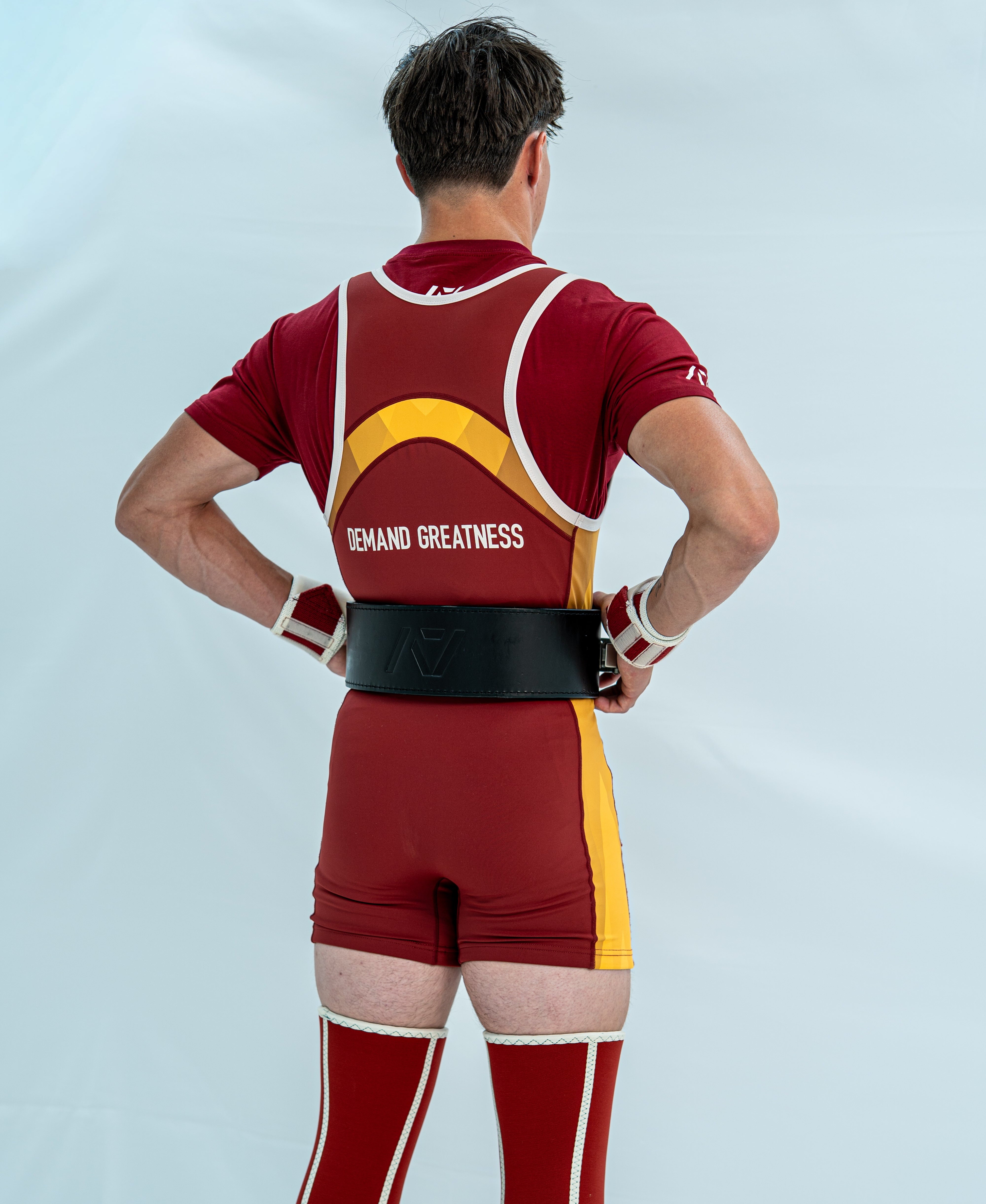 Male model wearing a red and yellow powerlifting singlet with 'Demand Greatness' on the back of the singlet on a light grey background