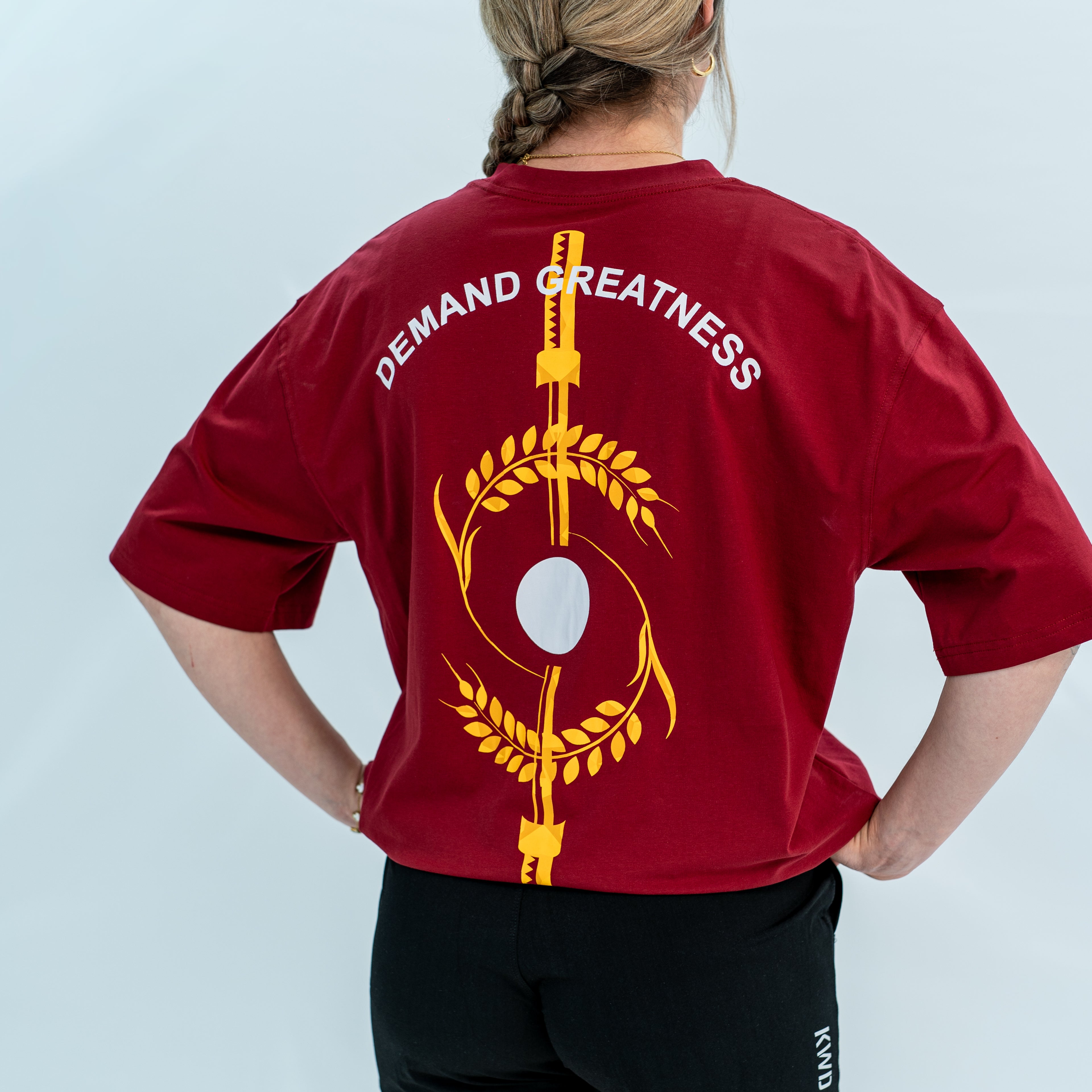 Female wearing a red unisex Over-Fit Shirt with 'Demand Greatness' text and design on the back, standing against a gray background.