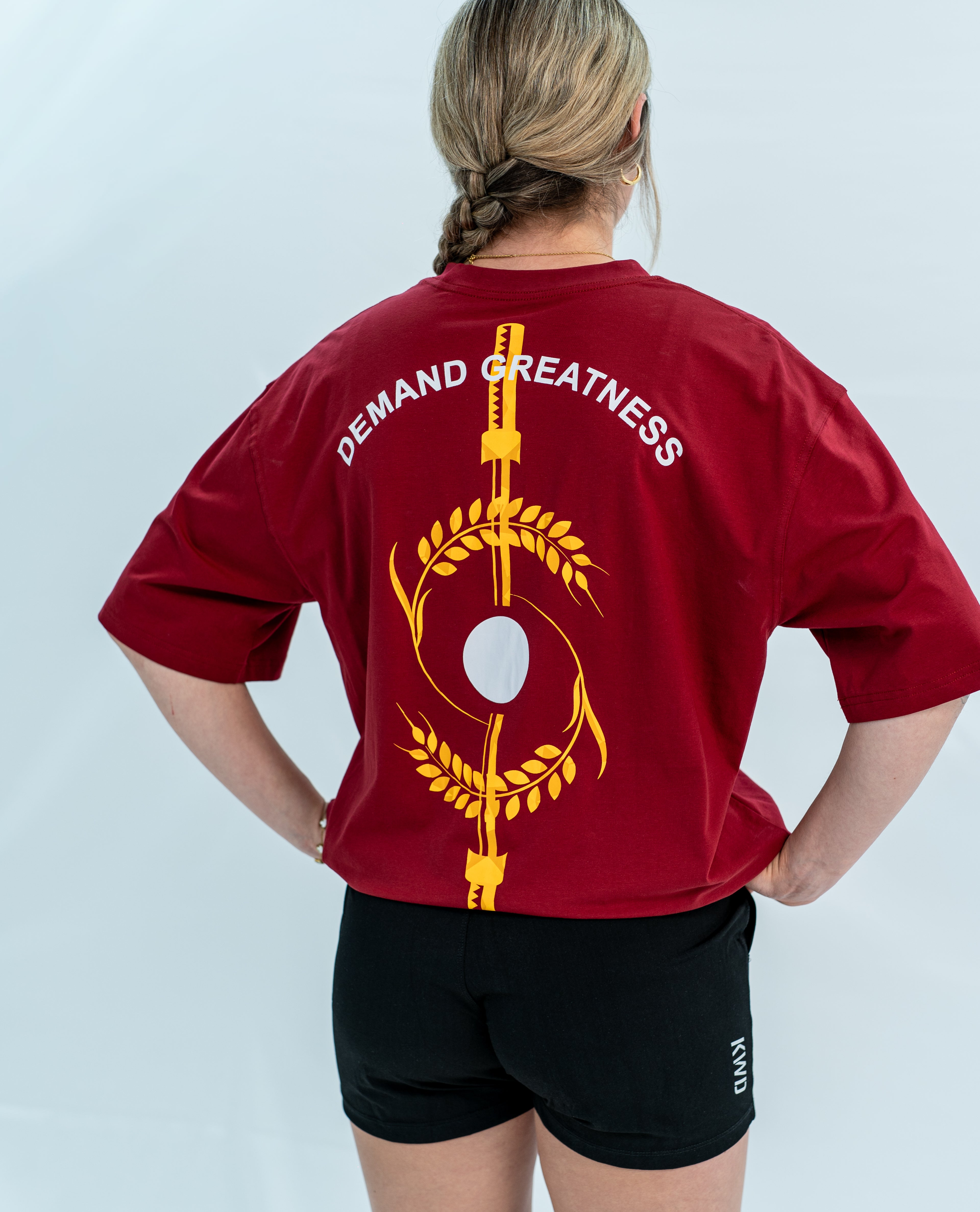 Female wearing a red unisex Over-Fit Shirt with 'Demand Greatness' text and design on the back, standing against a gray background.