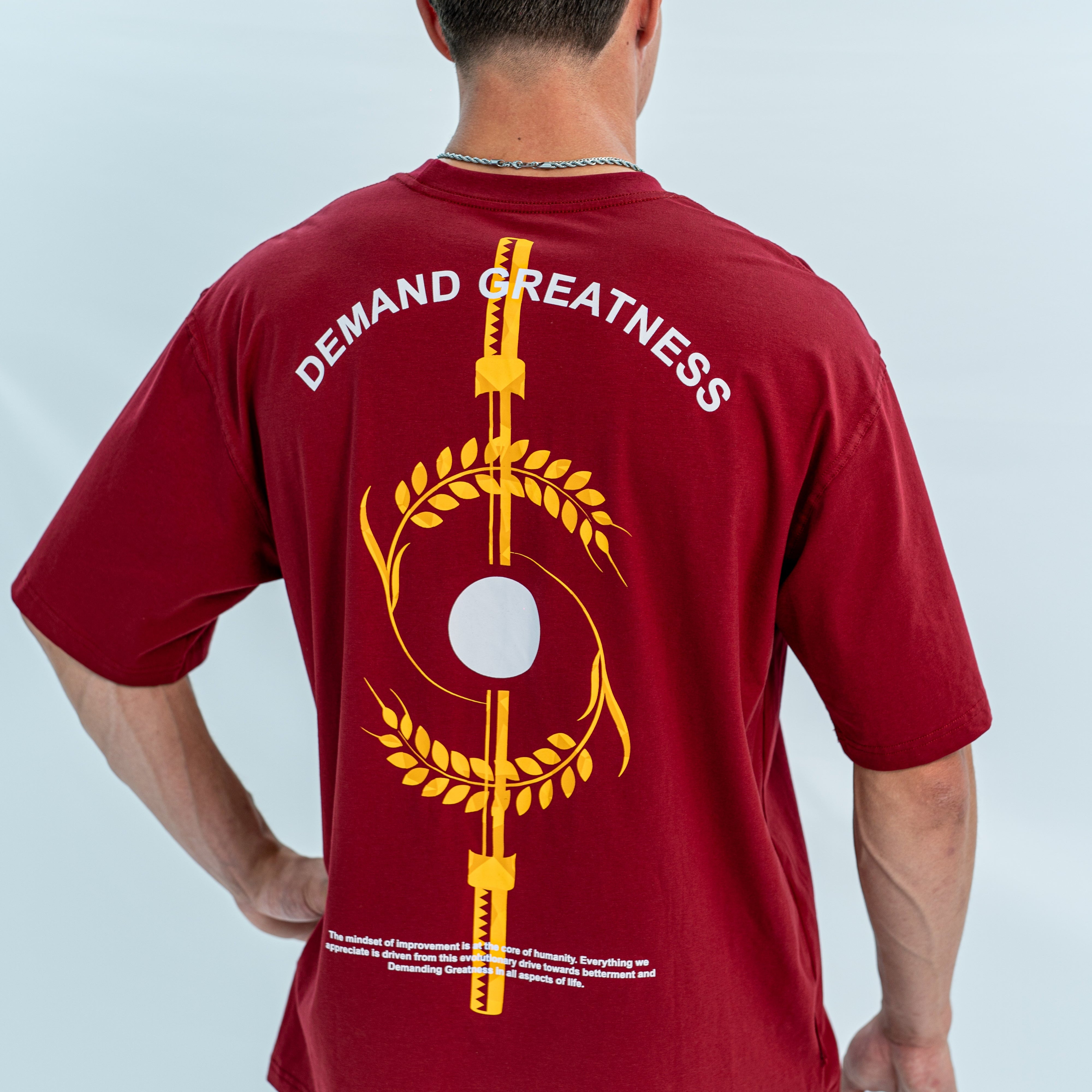 Red over-fit shirt with 'Demand Greatness' text and graphic on the back with equinox design, worn by a person against a light gray background