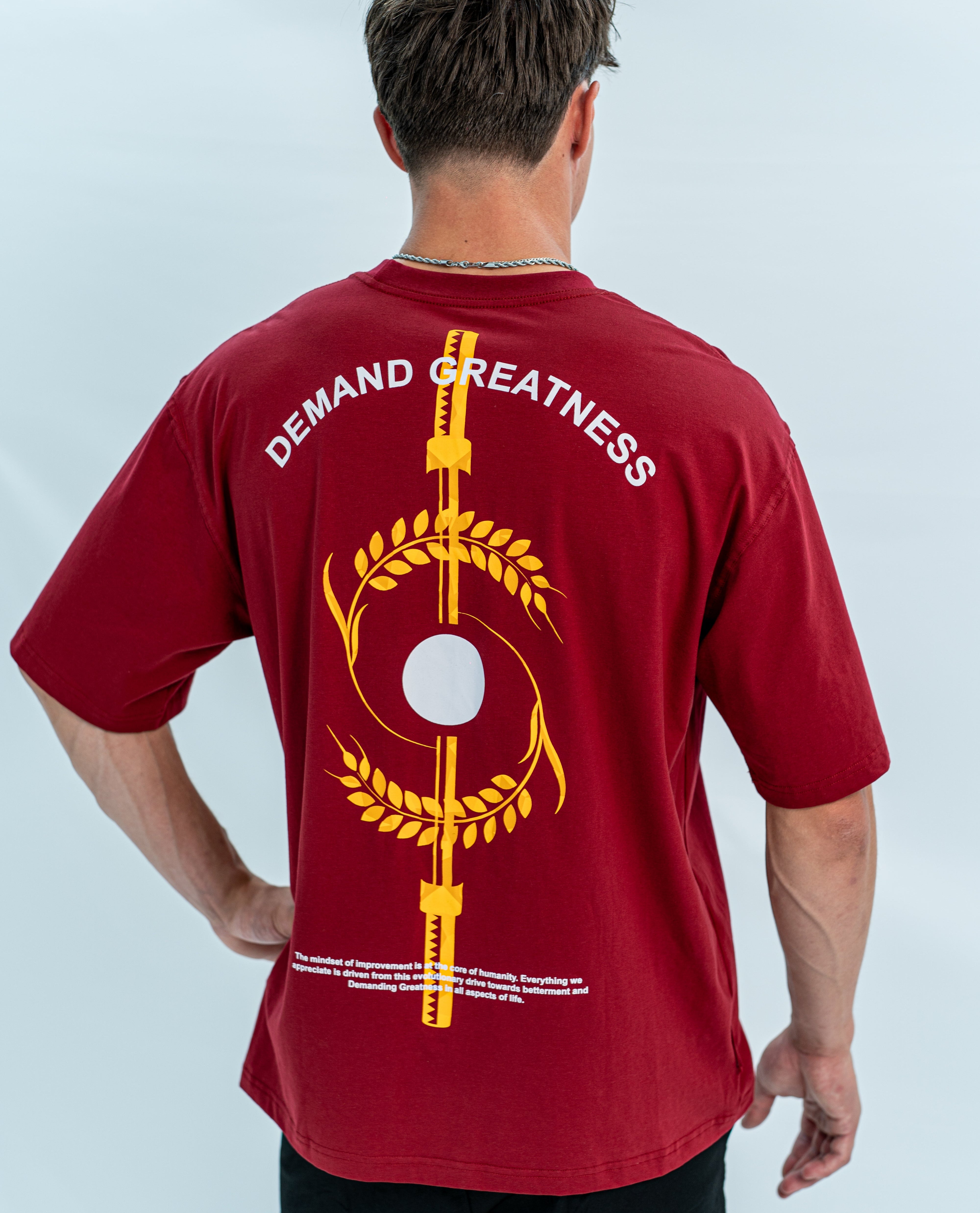 Red over-fit shirt with 'Demand Greatness' text and graphic on the back with equinox design, worn by a person against a light gray background