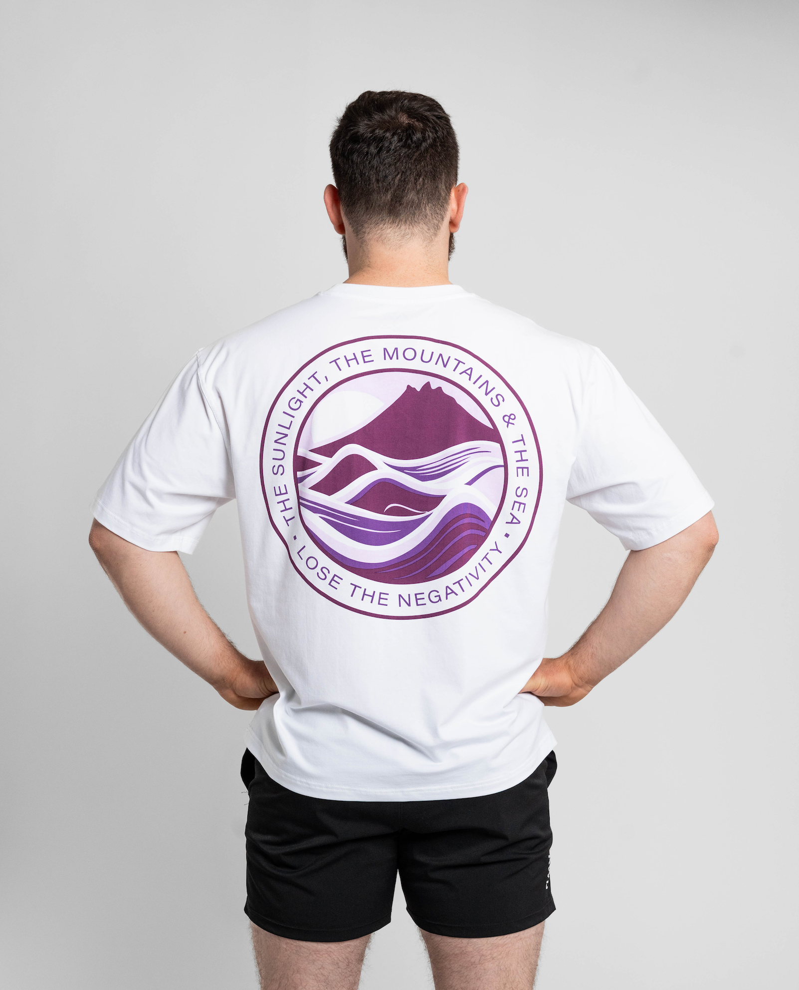 Person wearing a white t-shirt with a purple logo on the back, standing against a plain background