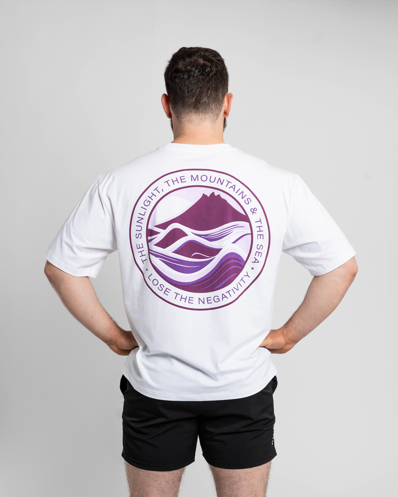 Person wearing a white t-shirt with a purple logo on the back, standing against a plain background