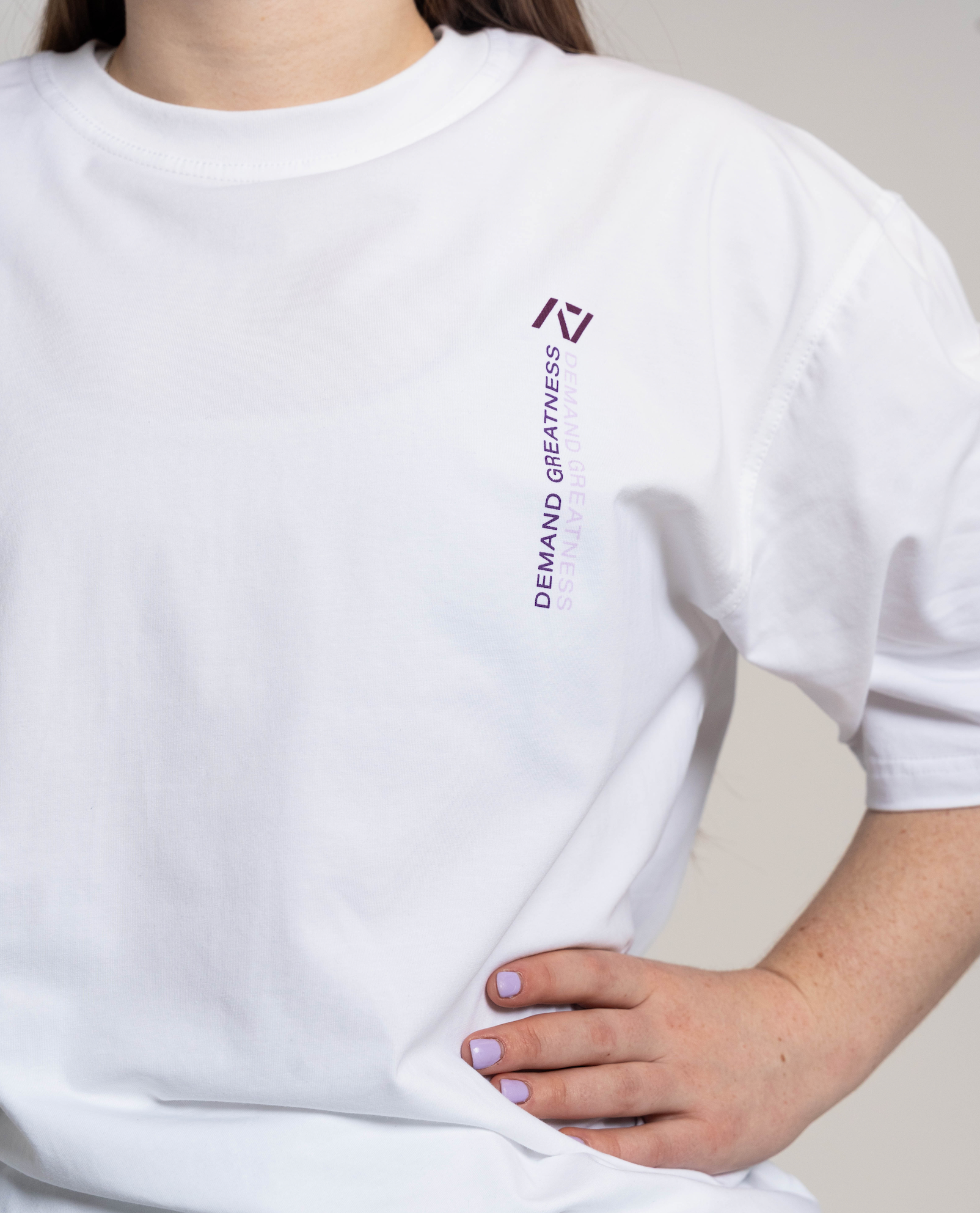White oversized t-shirt with a logo and text worn by a person, on a neutral background