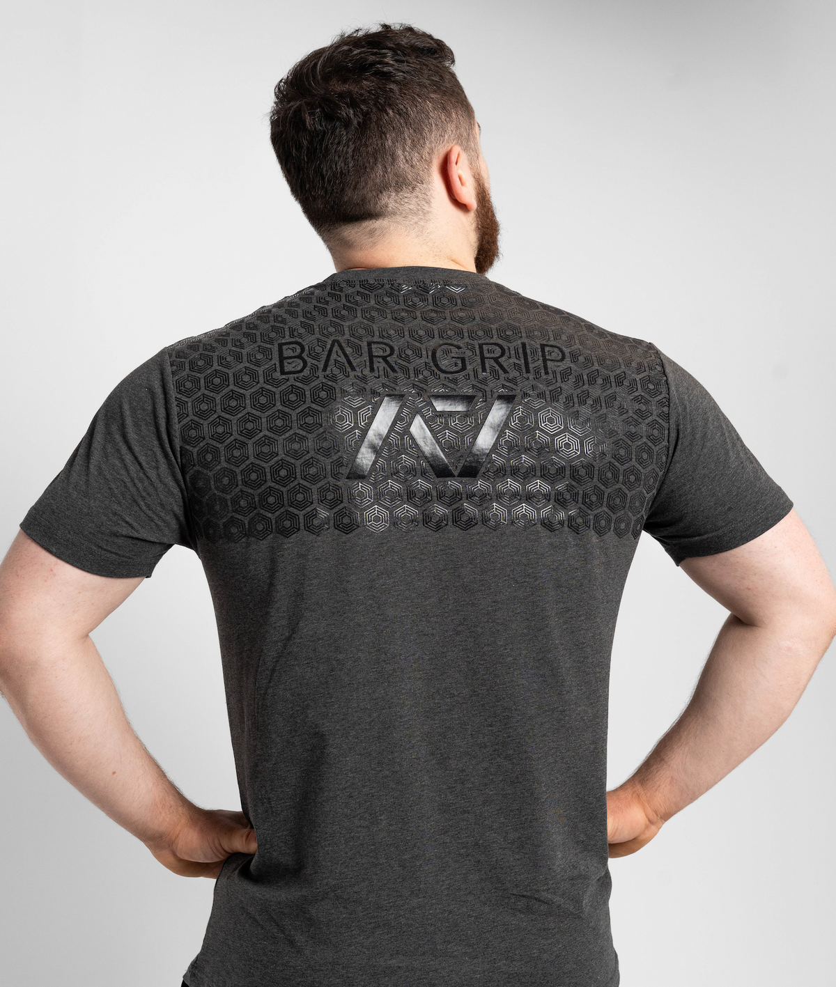 Man wearing a dark gray t-shirt with a7 bar grip on the back against a light gray background