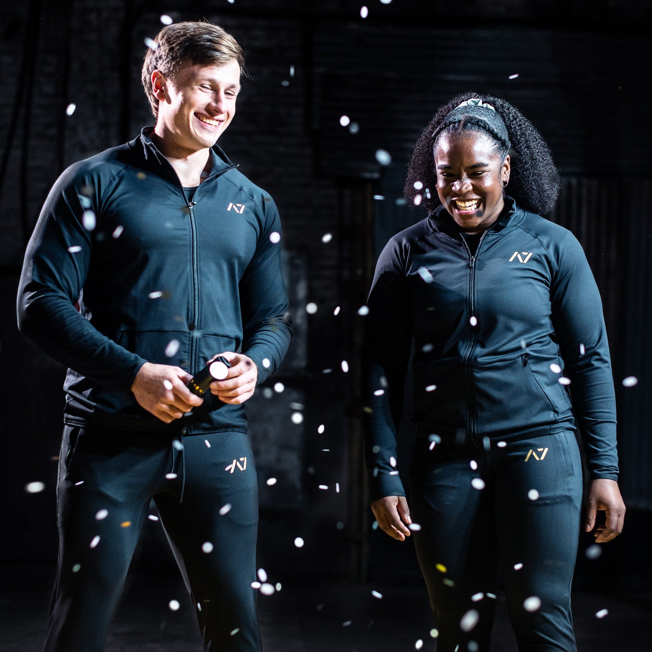 Male and female athletes wearing the Gold Standard Defy Joggers, standing side by side to highlight the unisex fit, tapered silhouette, and gold silicone A7 branding.