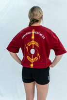Female wearing a red unisex Over-Fit Shirt with 'Demand Greatness' text and design on the back, standing against a gray background.